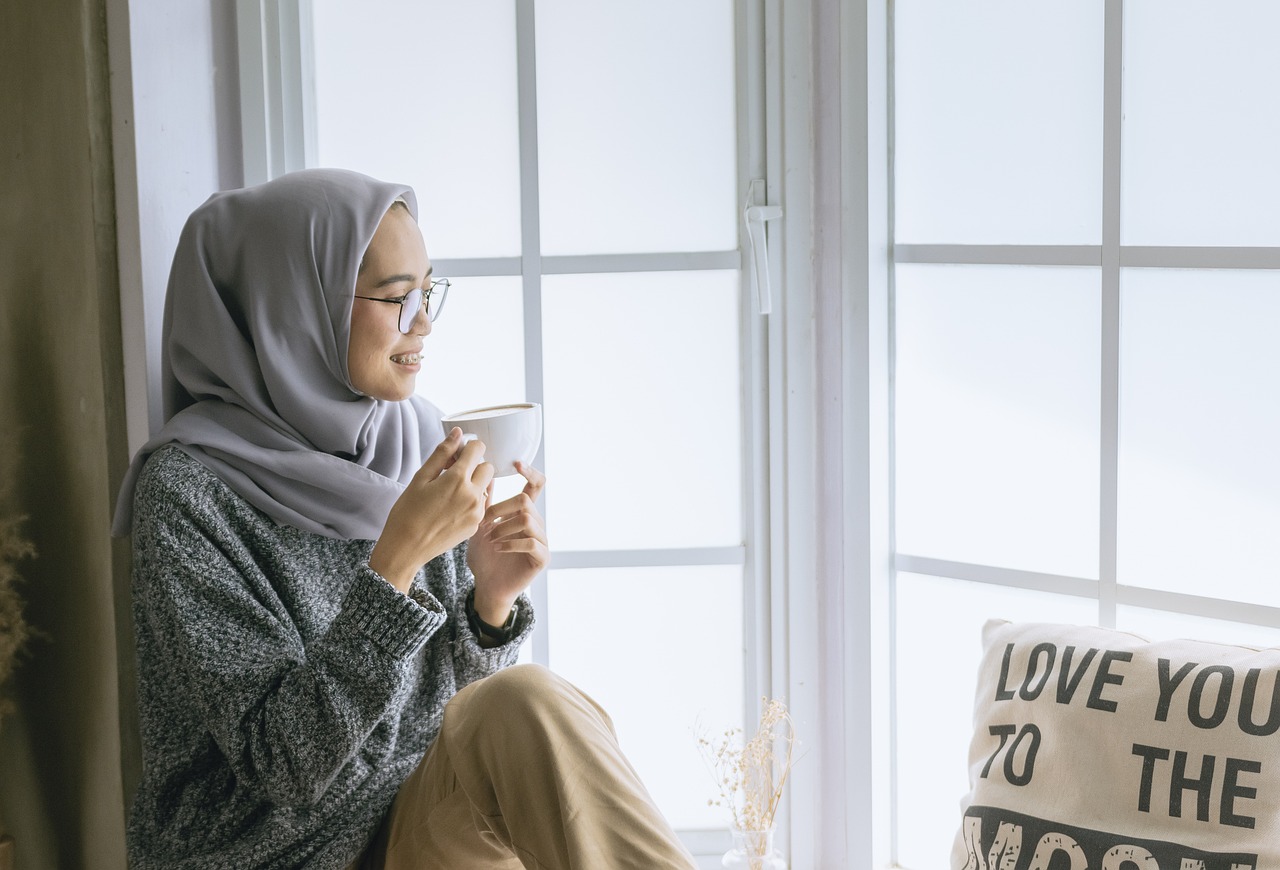 Woman wearing a hijab, drinking a cup of tea, sitting on a ledge looking out a window with a cushion that says 'Love you to the Moon'